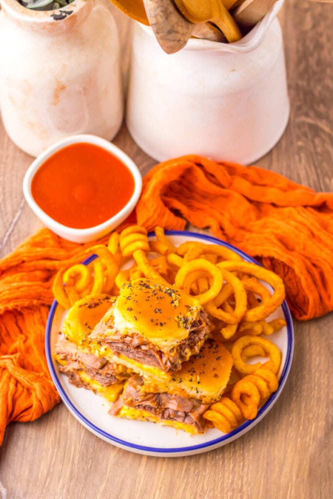 A plate with cheesy pulled pork sandwiches and curly fries sits on a wooden table next to a bowl of red dipping sauce, orange cloth, and ceramic containers with wooden utensils.
