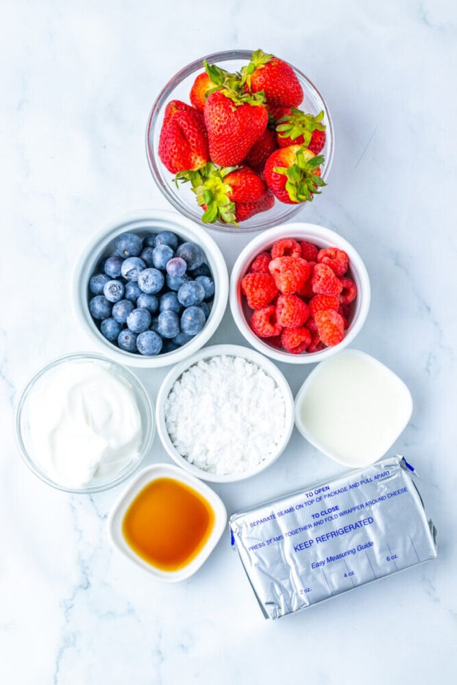 Six bowls and a package containing strawberries, raspberries, blueberries, powdered sugar, heavy cream, vanilla extract, Greek yogurt, and cream cheese on a white surface.