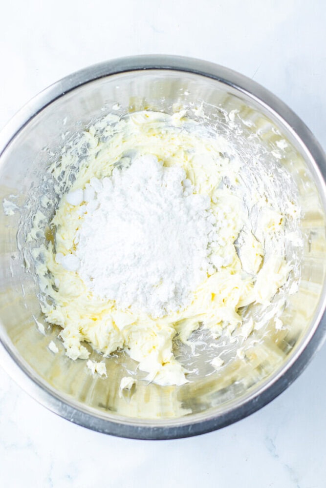 A metal mixing bowl containing creamed butter with a mound of powdered sugar on top, ready to be mixed together.