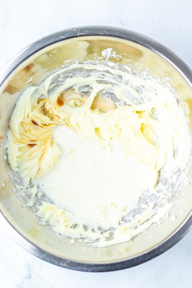 A metal mixing bowl containing creamed butter and sugar with some vanilla extract and cream added, ready to be mixed.