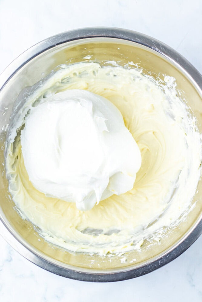 A metal mixing bowl containing whipped cream cheese mixture, with a dollop of whipped cream on top, viewed from above.