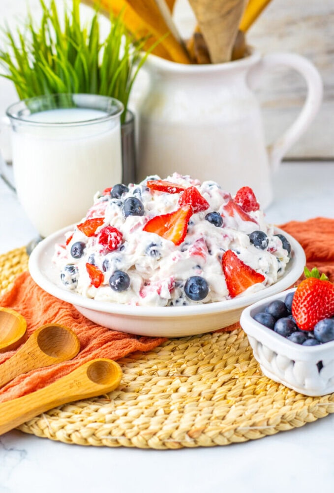 A bowl of creamy fruit salad with strawberries and blueberries sits on a woven mat, next to a glass of milk, wooden spoons, and a small bowl of blueberries.