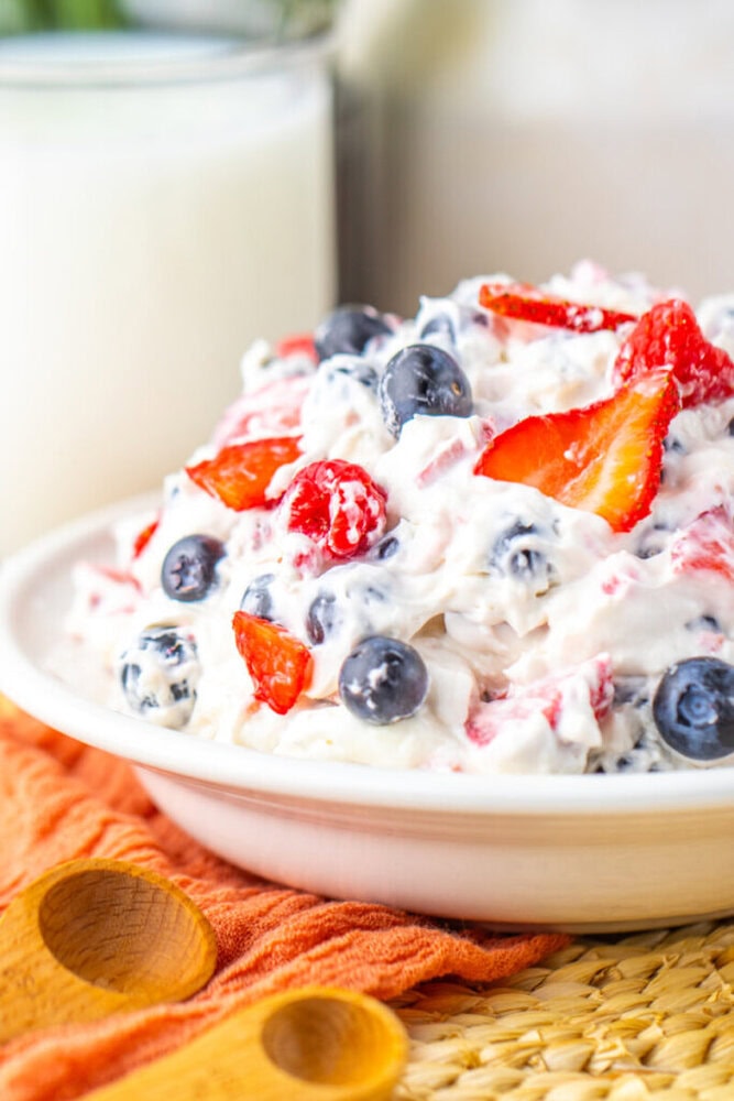 A bowl of creamy fruit salad with blueberries, strawberries, and raspberries, placed on an orange cloth next to wooden spoons and a glass of milk.
