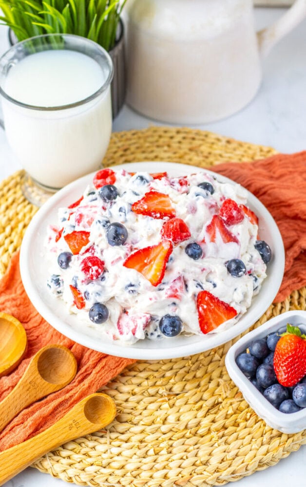 A plate of creamy fruit salad with strawberries and blueberries sits on a woven mat beside a bowl of blueberries, a glass of milk, and wooden measuring spoons.