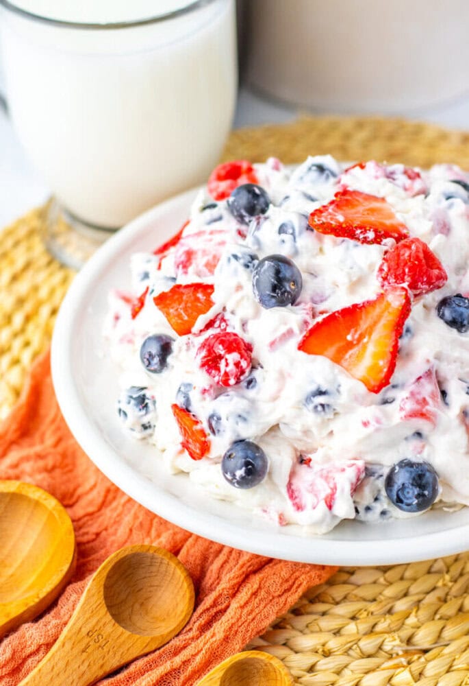 A bowl of creamy fruit salad with strawberries and blueberries sits on a woven placemat next to a glass of milk, an orange napkin, and wooden measuring spoons.
