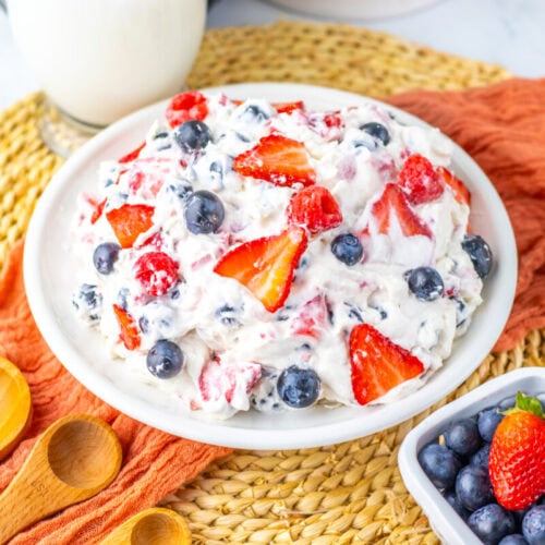 A bowl of creamy fruit salad with strawberries and blueberries on a woven placemat, next to wooden spoons and a small bowl of blueberries and a strawberry.