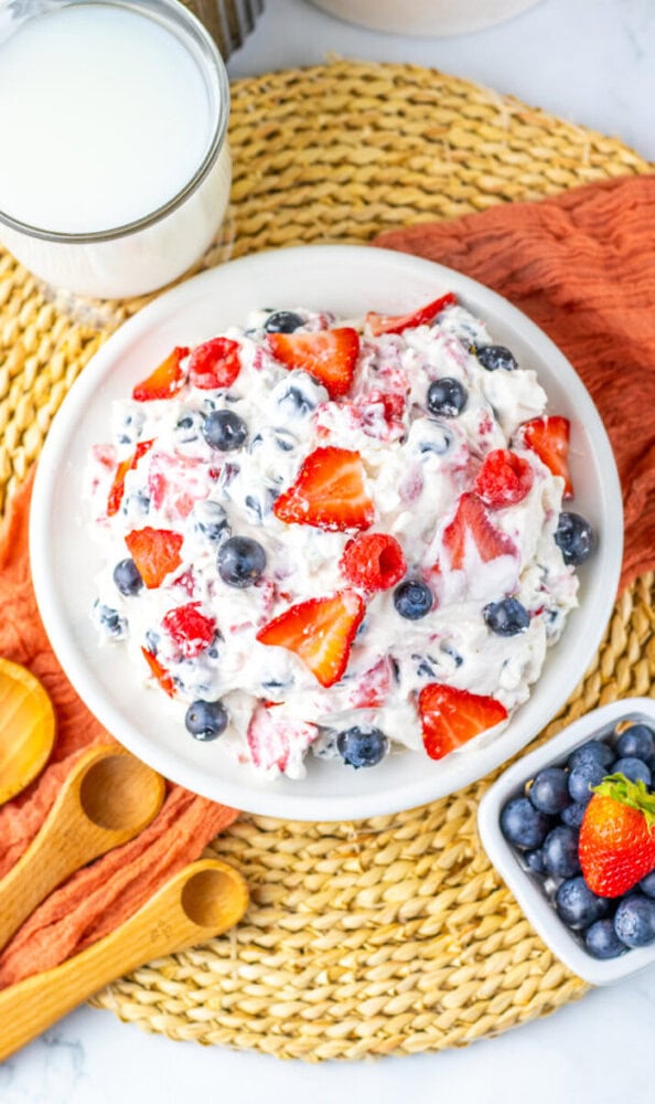 A bowl of mixed berry salad with whipped cream, featuring strawberries and blueberries, sits on a woven placemat beside a small dish of blueberries, wooden spoons, and a glass of milk.