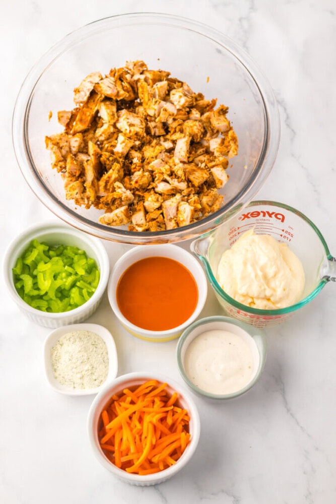 A glass bowl of chopped cooked chicken surrounded by bowls of sliced celery, shredded carrots, ranch seasoning, hot sauce, mayonnaise, and ranch dressing on a marble surface.