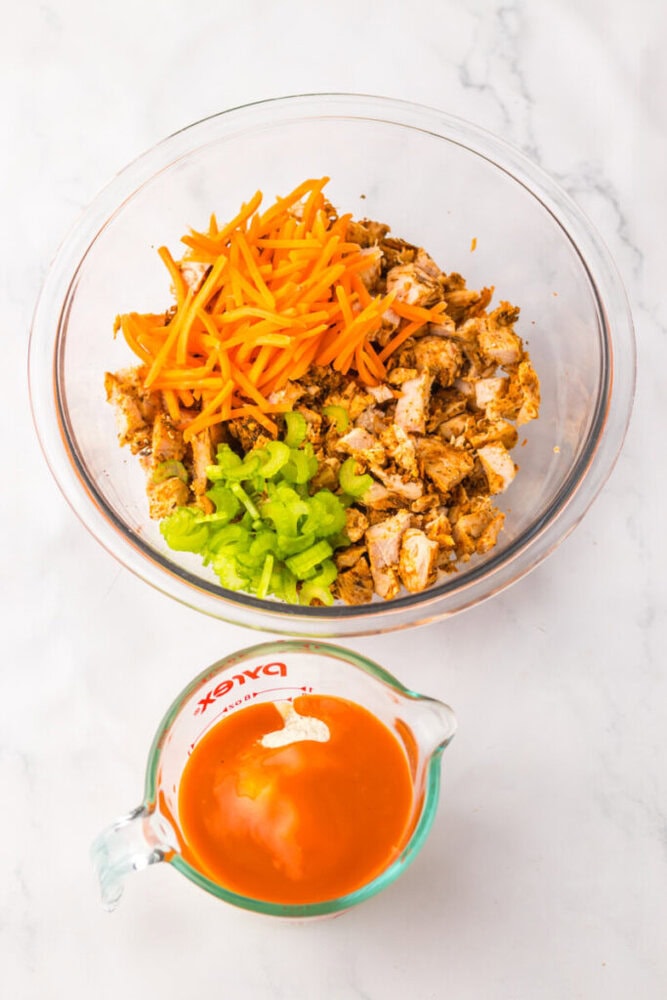 A glass bowl with chopped cooked chicken, shredded carrots, and celery sticks, next to a measuring cup containing buffalo sauce on a white surface.