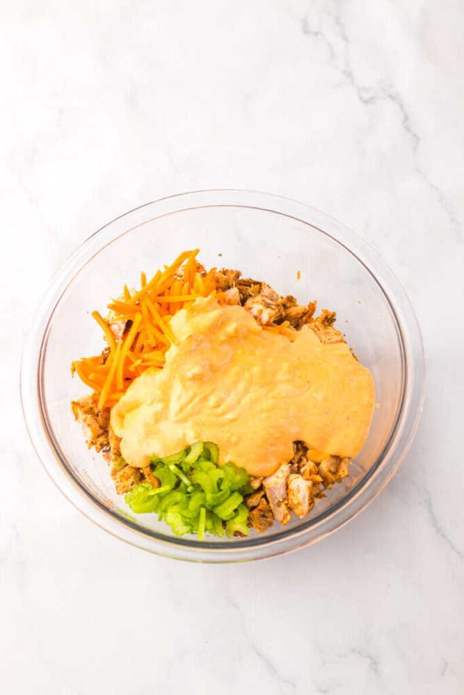 A glass bowl containing shredded chicken, grated carrots, sliced green bell pepper, and a layer of dough or batter on top, set on a white marble surface.