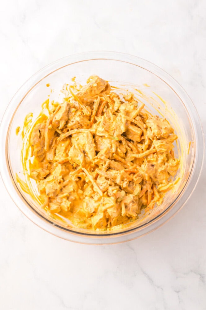 A glass bowl containing a mixture of shredded ingredients coated in a creamy yellow sauce sits on a white marble surface.