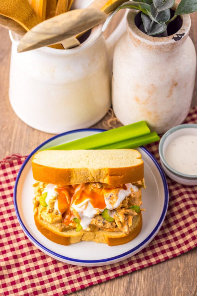 A plate with a buffalo chicken sandwich topped with celery, ranch dressing, and hot sauce, next to a small bowl of ranch and a celery stick, on a checkered napkin.