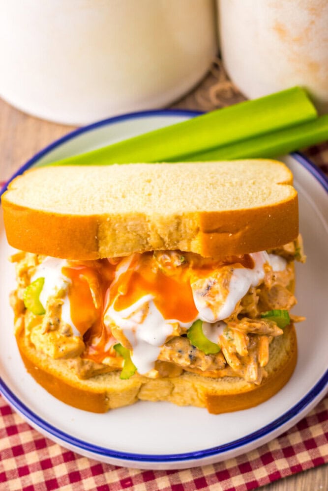 A buffalo chicken sandwich topped with ranch dressing and served with celery sticks on a white plate.