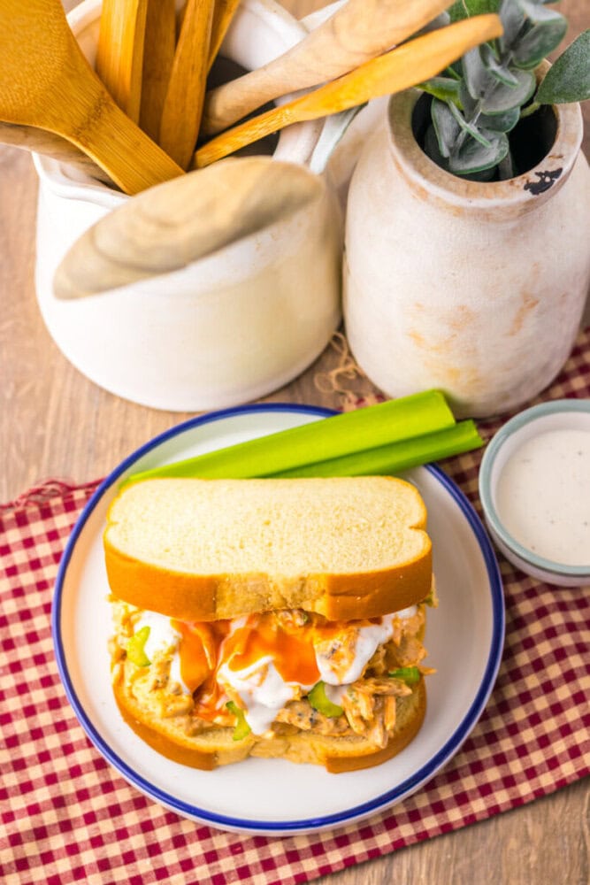 A buffalo chicken sandwich with celery sticks and a small bowl of ranch dressing on a plate, placed on a red checkered napkin. Wooden utensils and a vase are in the background.