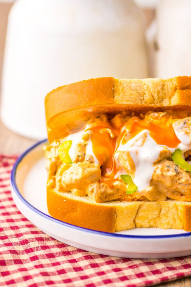 A close-up of a chicken salad sandwich with buffalo sauce and ranch dressing on thick white bread, placed on a white plate with a blue rim.
