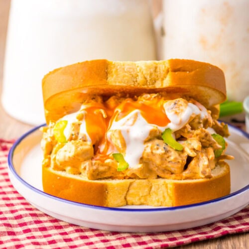 A chicken sandwich on thick white bread is topped with buffalo sauce and ranch dressing, served on a white plate with a red checkered napkin underneath.