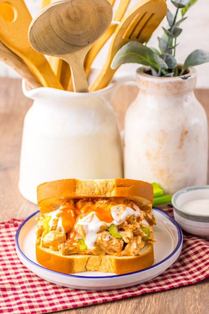A buffalo chicken sandwich with lettuce, ranch, and buffalo sauce is served on a plate, with wooden utensils and a ceramic vase in the background.