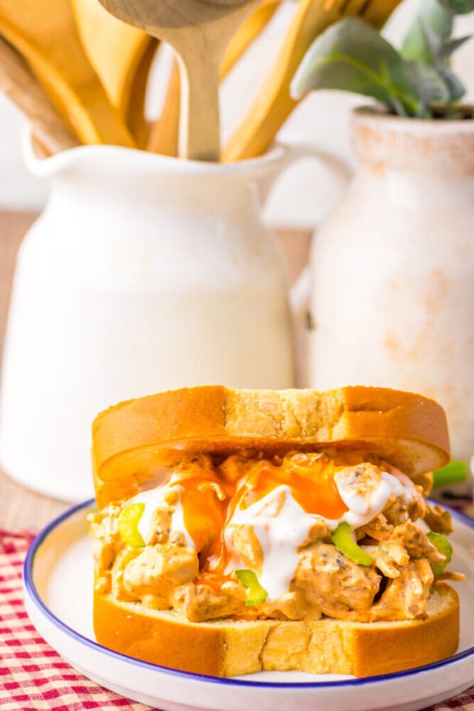 A chicken salad sandwich with hot sauce and ranch dressing on white bread sits on a plate, with ceramic jars and wooden utensils in the background.