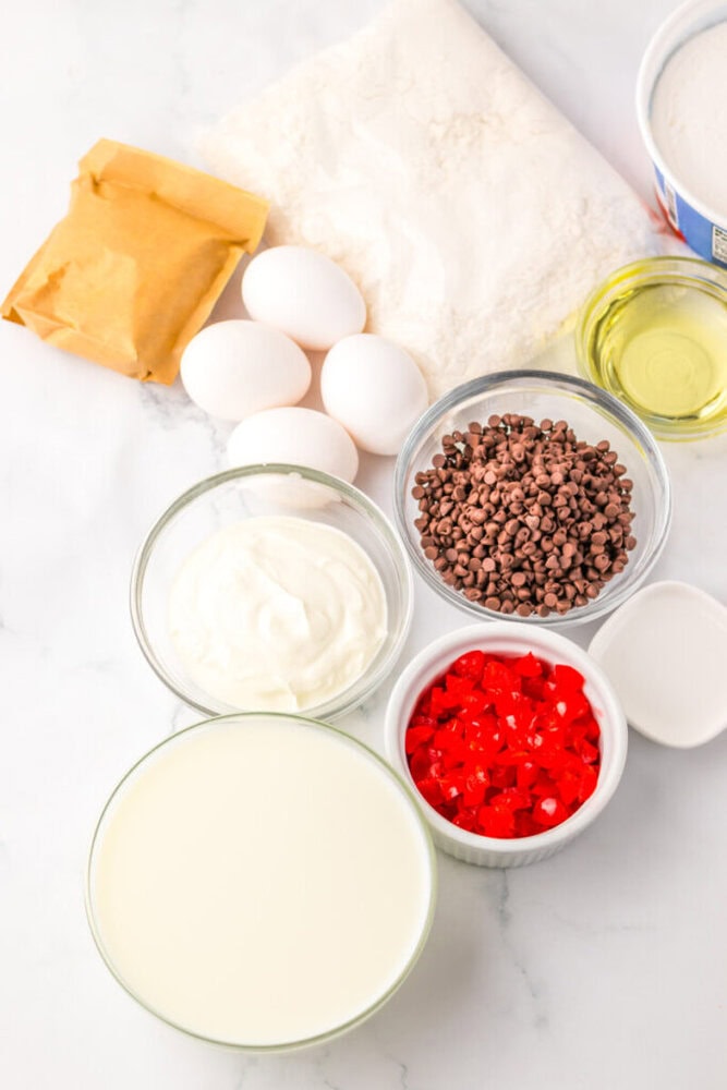 Assorted baking ingredients on a white surface, including eggs, flour, oil, cream, milk, chocolate chips, and chopped red candied cherries.