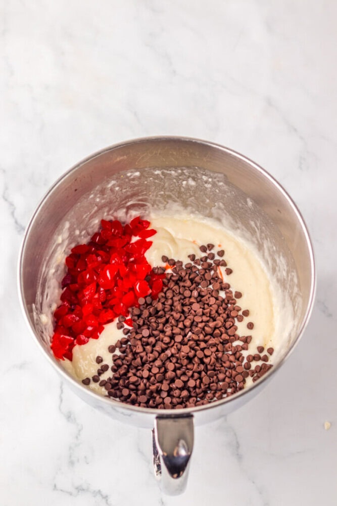 A metal mixing bowl with cake batter, mini chocolate chips, and chopped red candied cherries on a white marble surface.