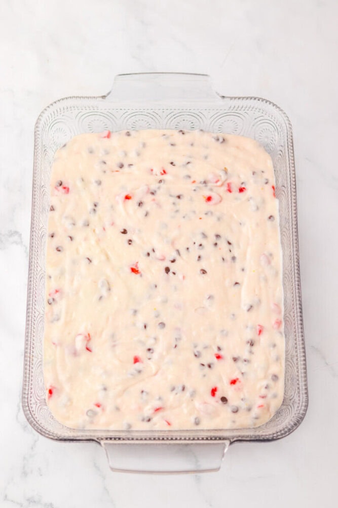 A glass baking dish filled with unbaked batter containing chocolate chips and red fruit pieces on a white surface.