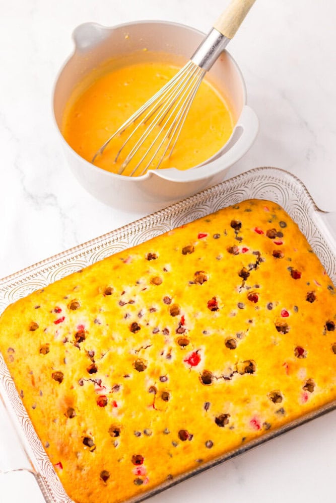 A baked sheet cake with visible berries and chocolate chips sits next to a mixing bowl filled with yellow batter and a whisk on a white surface.