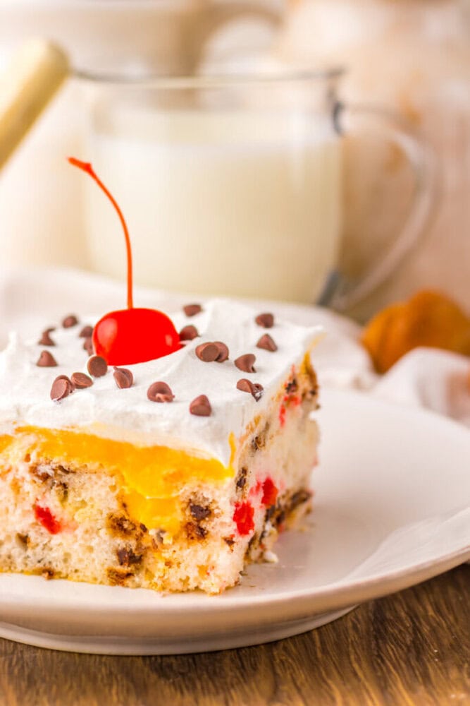 A slice of cake with white frosting, yellow filling, chocolate chips, and a cherry on top, on a white plate with a glass of milk in the background.