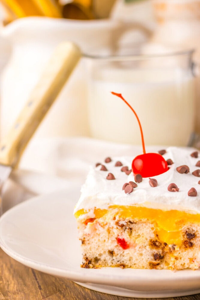 A slice of cake with white frosting, chocolate chips, yellow filling, and a cherry on top, served on a white plate with a glass of milk in the background.