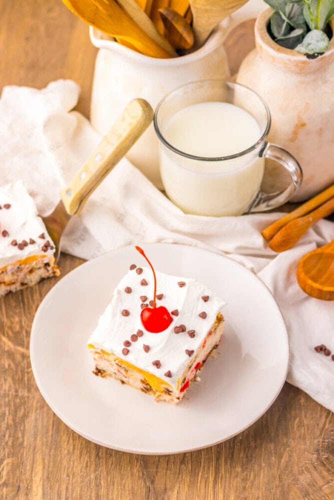 A slice of layered dessert topped with whipped cream, chocolate chips, and a maraschino cherry on a white plate next to a glass mug of milk and wooden utensils.