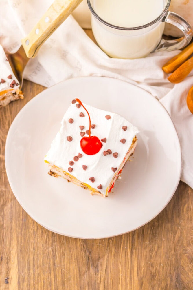 A square slice of dessert topped with whipped cream, mini chocolate chips, and a cherry, served on a white plate with a glass of milk nearby.