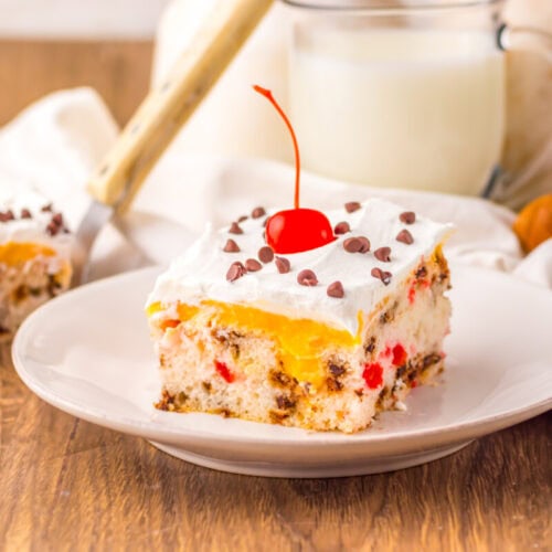 A slice of white cake with whipped topping, chocolate chips, a cherry, and pieces of fruit sits on a white plate with a glass of milk in the background.