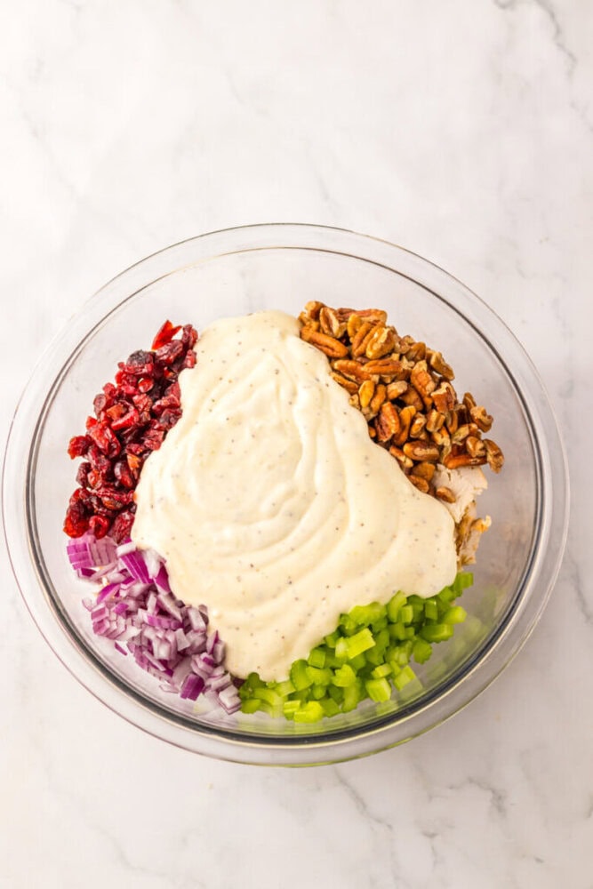 A glass bowl containing diced celery, chopped red onion, pecans, dried cranberries, and chicken, topped with a creamy dressing, on a white marble surface.