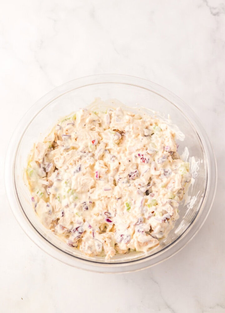 A glass bowl filled with a creamy mixture containing chopped celery, onions, and what appears to be chicken salad on a white surface.
