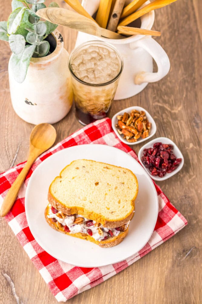 A chicken salad sandwich on a white plate with a red checkered napkin, served with iced tea, pecans, dried cranberries, and a wooden spoon on a wooden table.