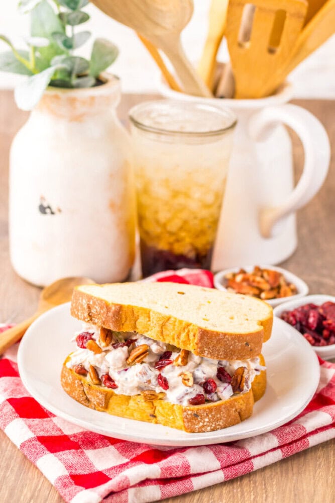 A chicken salad sandwich with pecans and cranberries sits on a white plate with a drink and small bowls of nuts and cranberries in the background.