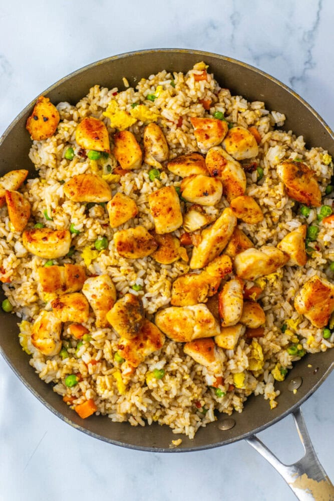 A skillet filled with fried rice, mixed vegetables, scrambled eggs, and pieces of cooked chicken on a marble surface.