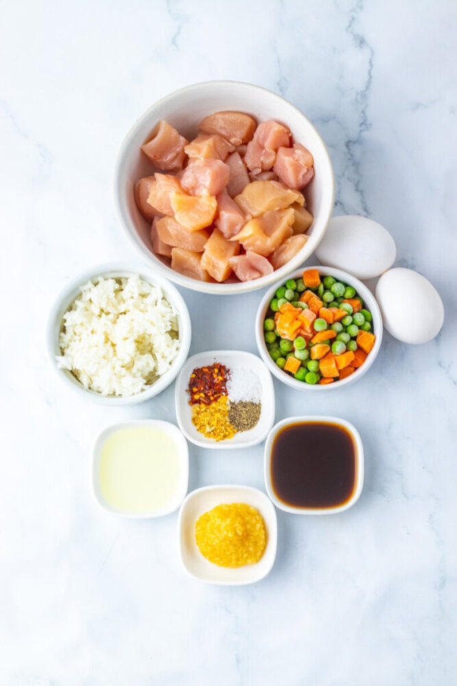 Bowls containing diced raw chicken, two eggs, mixed vegetables, riced cauliflower, oil, minced garlic, soy sauce, and spices arranged on a white surface.