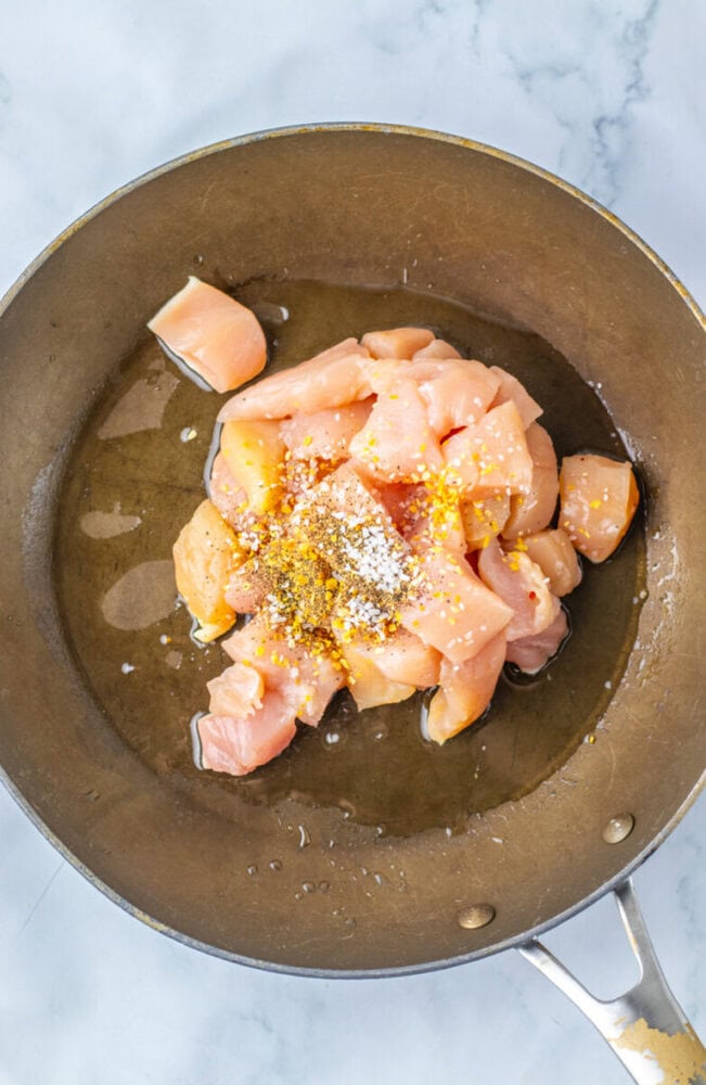 Raw cubed chicken pieces with oil and seasonings are in a frying pan, ready to be cooked on a marble countertop background.