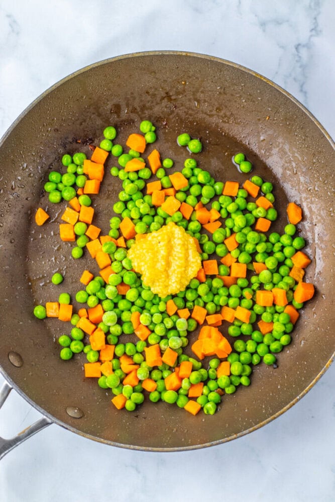 Diced carrots, green peas, and a dollop of minced garlic are being sautéed in a large pan on a marble surface.