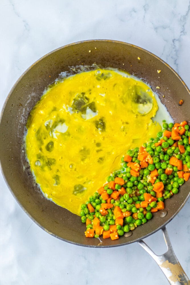 A frying pan with partly cooked scrambled eggs on one side and a mixture of peas and diced carrots on the other, seen from above.