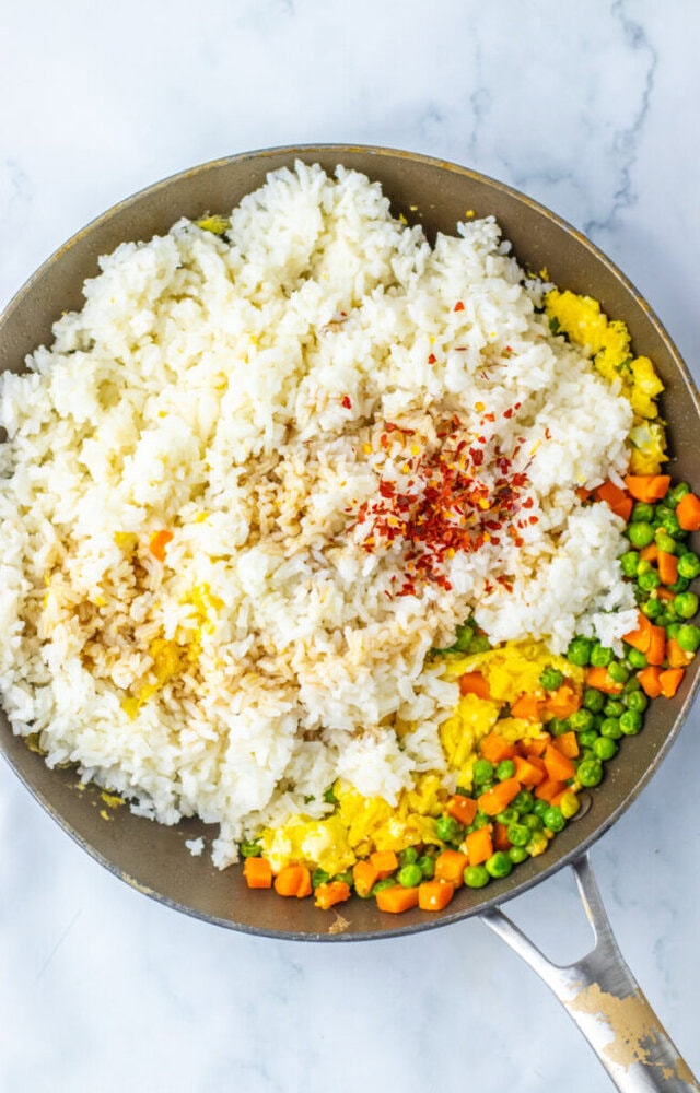 A skillet containing white rice, peas, carrots, scrambled eggs, soy sauce, and red pepper flakes, ready to be mixed for fried rice.