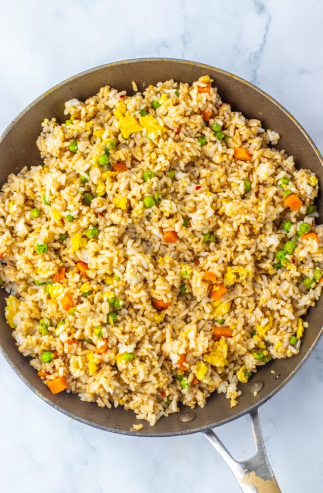 A skillet filled with fried rice containing peas, diced carrots, scrambled egg, and rice, mixed with soy sauce on a marble surface.
