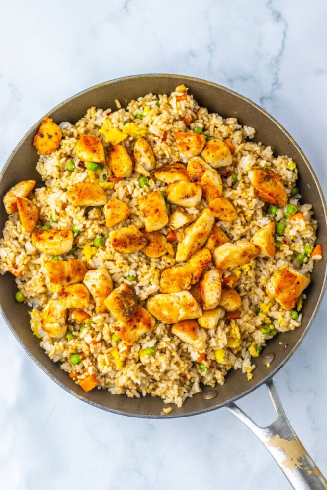 A skillet filled with fried rice, mixed vegetables, and pieces of cooked, seasoned chicken on a marble surface.