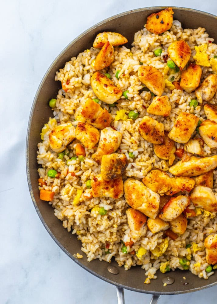 A skillet filled with fried rice, mixed vegetables, scrambled eggs, and seasoned chicken pieces on a light surface.
