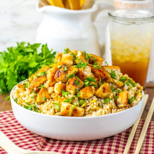 A bowl of rice topped with seasoned grilled chicken pieces, garnished with herbs, sits on a table with a wooden spoon, chopsticks, and a glass of iced drink in the background.