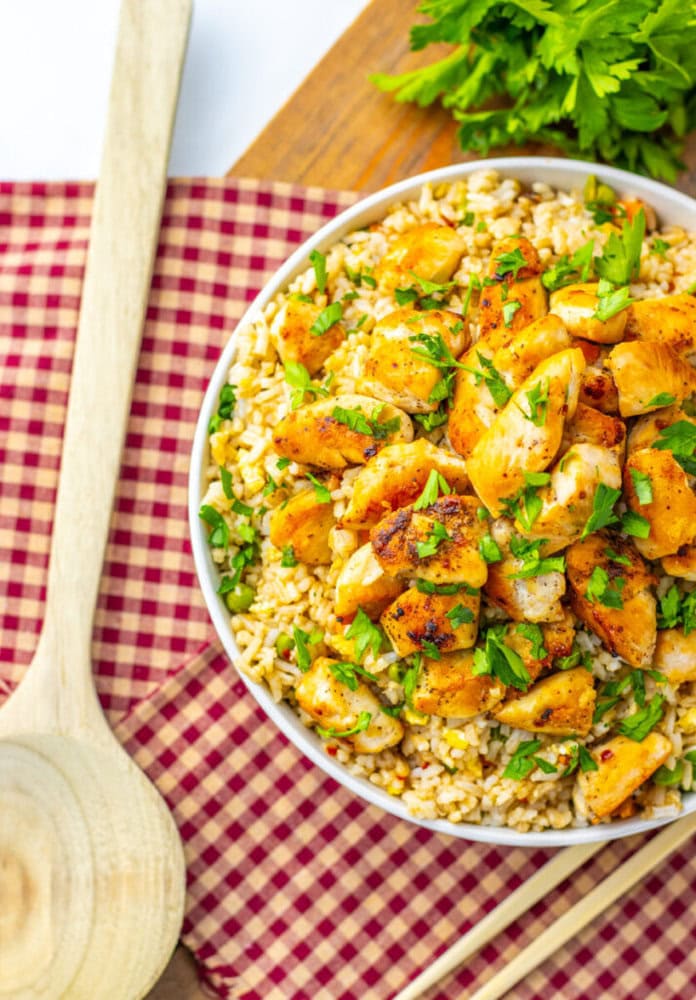 A bowl of seasoned, cooked chicken pieces on brown rice, garnished with chopped parsley, placed on a red checkered cloth beside a wooden spoon and fresh herbs.