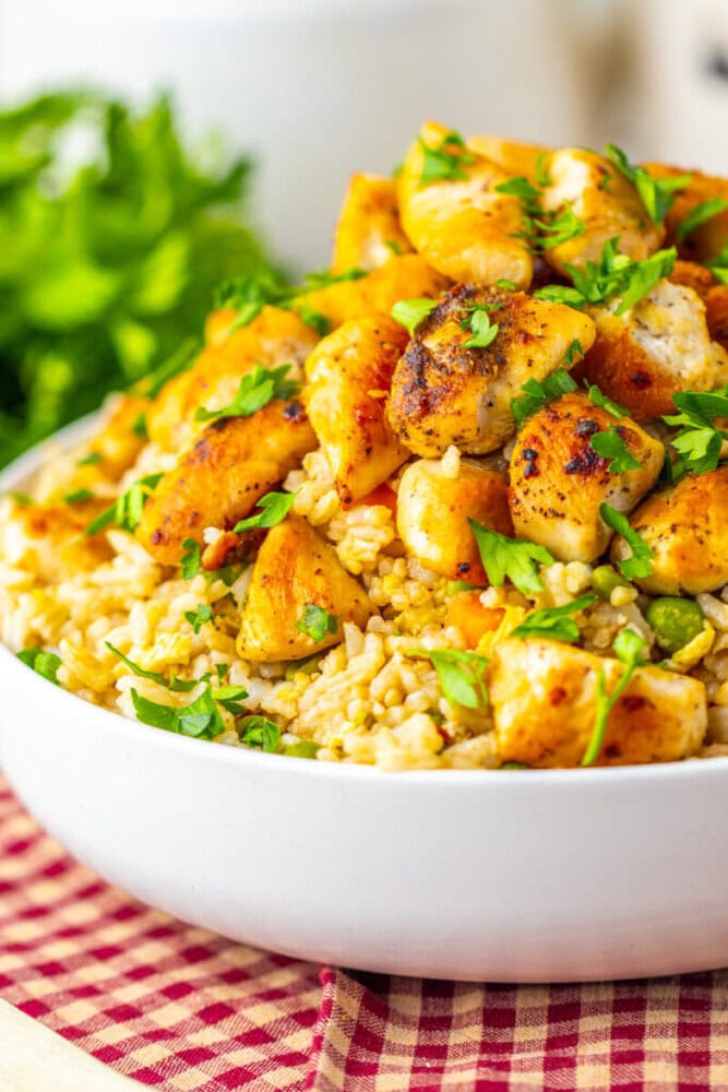 A bowl of cooked brown rice topped with grilled, seasoned chicken pieces and garnished with chopped fresh herbs.