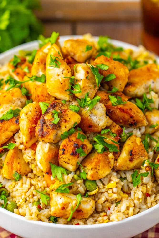A bowl of cooked rice topped with seasoned, grilled chicken pieces and garnished with chopped fresh herbs.