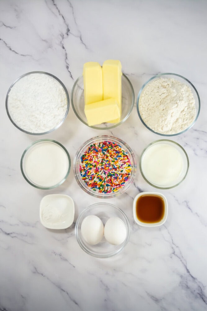Assorted baking ingredients in glass bowls on a marble surface, including butter, flour, sugar, milk, eggs, rainbow sprinkles, baking powder, and vanilla extract.