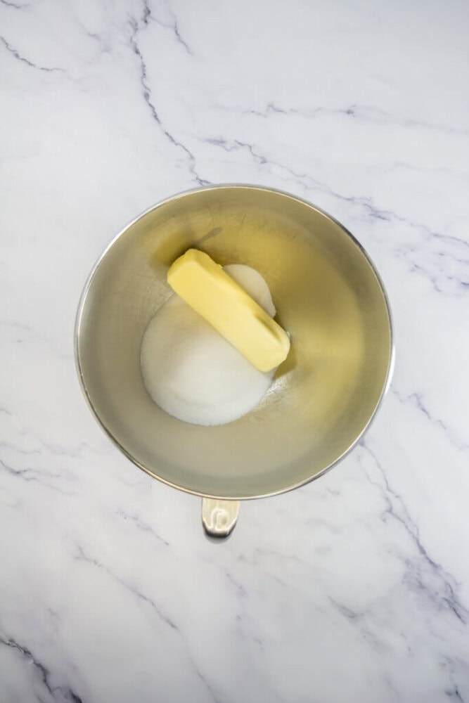 A stick of butter and a pile of granulated sugar are in a metal mixing bowl on a white marble countertop.